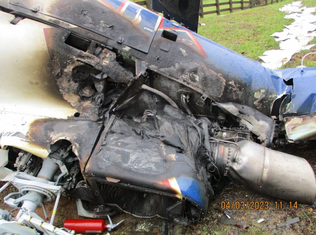 Close-up of the helicopter’s burned engine compartment and fuselage, displaying extensive fire damage, melted components, and charred paint on the blue and white airframe.